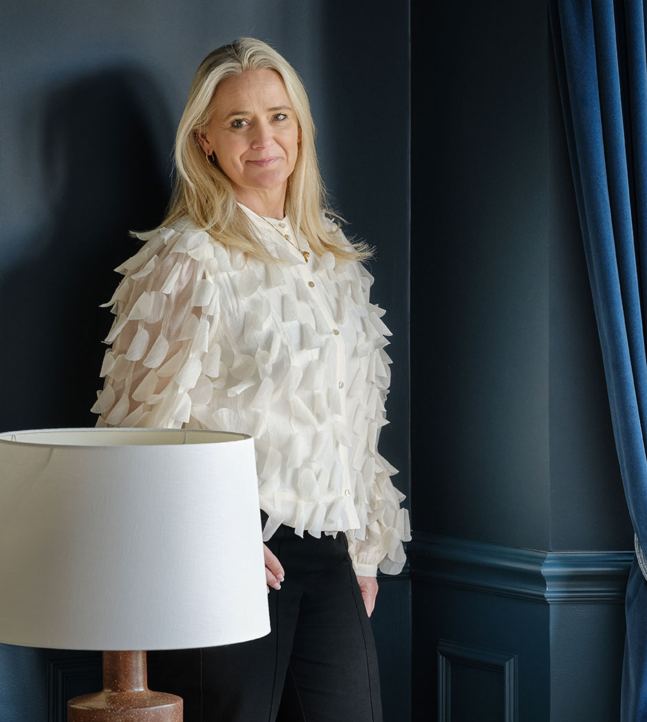 Elie Jones, Interior Designer standing in a luxurious living room featuring dark blue walls, velvet curtains, and chic furnishings